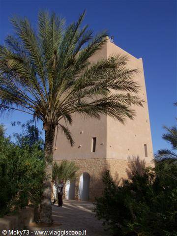 Rough mud-brick tower with palm tree and person near arched entrance, clear blue sky