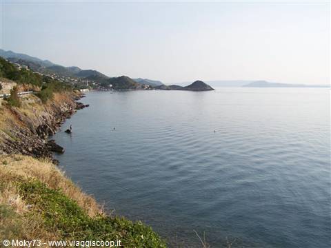 Rocky shoreline with mediterranean shrubs on calm sea, village and hills behind