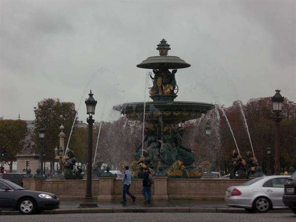 Monumental green and gold bronze fountain, pedestrians and cars under grey Paris sky