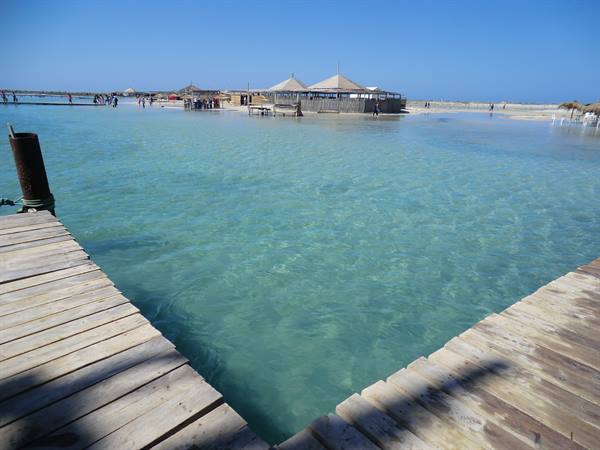 Wooden walkway over turquoise water, tents and visitors on the beach