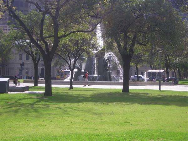 Sculptural fountain among trees in a park, passerby photographing on green lawn