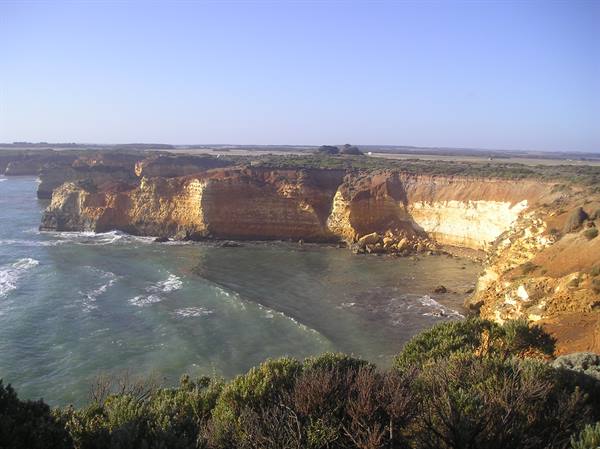 Ochre limestone cliffs over turquoise ocean, shrubs in foreground, morning light