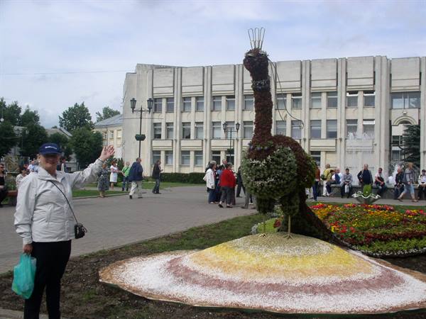 Woman waves beside a flower peacock sculpture on a town square