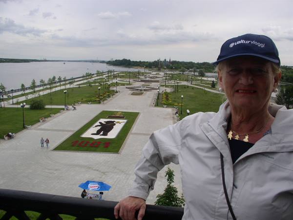 Woman in blue cap on terrace above park with river and bear-shaped flowerbed