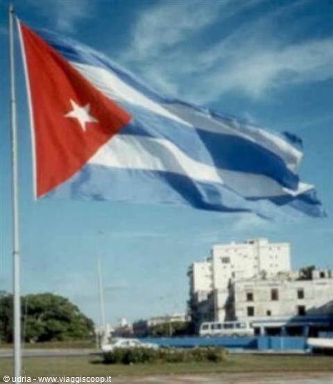 Cuban flag waving on metal pole, white buildings and bus in background