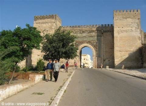 Massive stone gate with crenellated towers, people walking along the road