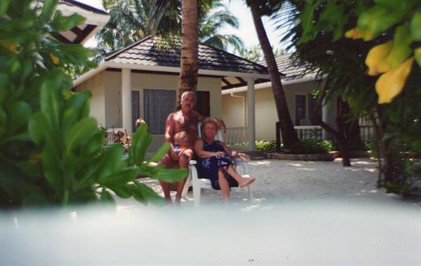 Couple on white lounge chairs among bungalows and palms, sandy tropical light