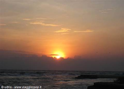 Orange sun sinking through clouds over choppy sea, dark rocks in foreground