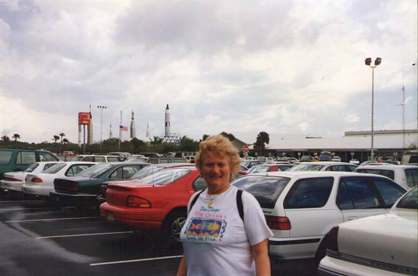 Smiling woman in Kennedy Space Center parking lot, rockets visible behind cars