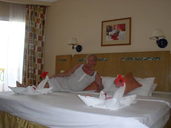 Woman in white dress sitting on hotel bed with towel swans and flowers