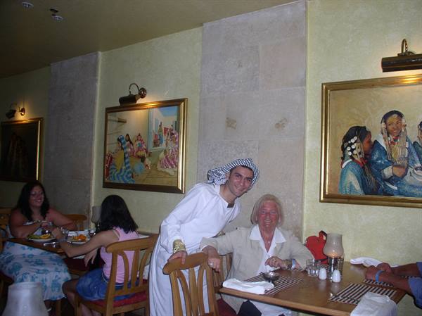 Waiter in white robe and keffiyeh beside a smiling woman at restaurant table