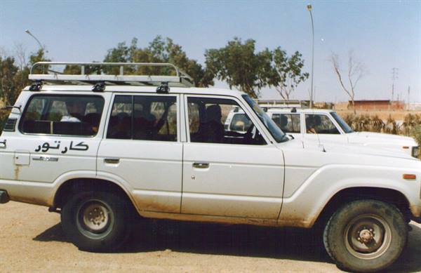 White Toyota Land Cruiser with Arabic lettering, sandy ground and trees behind