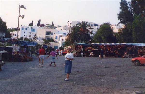 Tourist poses in square with market stalls, white houses and palms behind
