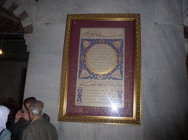 Framed Arabic calligraphy panel on marble wall, visitors photographed beside it