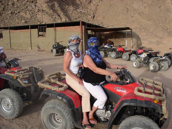 Two women wearing keffiyeh scarves sit on a red quad bike in the desert