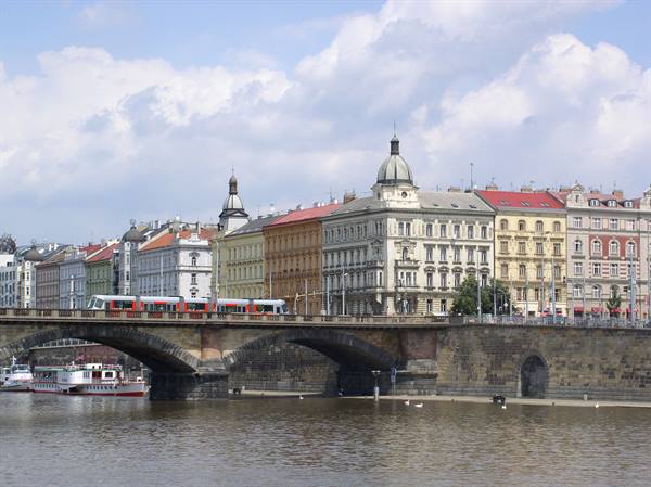 Red tram crossing Prague bridge over the Vltava, historic colorful buildings behind