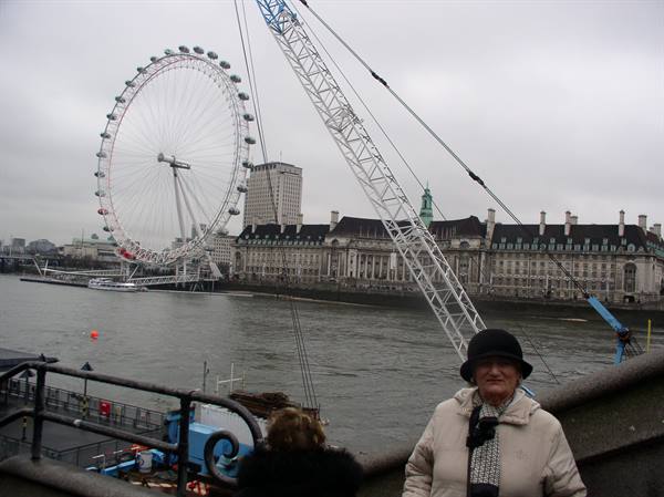 Woman in black hat near a crane, on the Thames, facing the London Eye