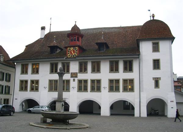 White town hall with red clock, arched loggia and fountain on cobbled square