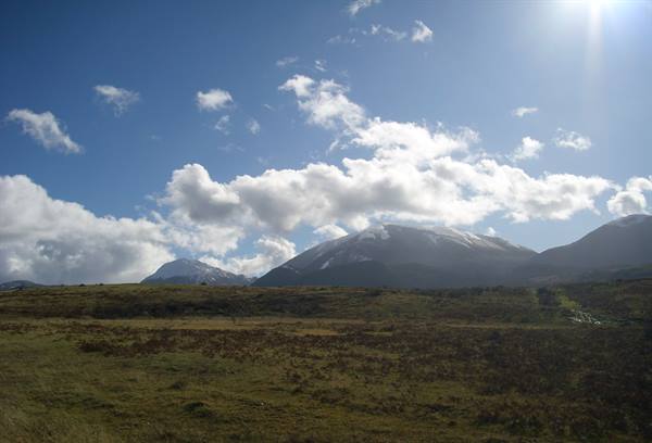 Snow-capped mountains over a grassy moorland, white clouds and sun