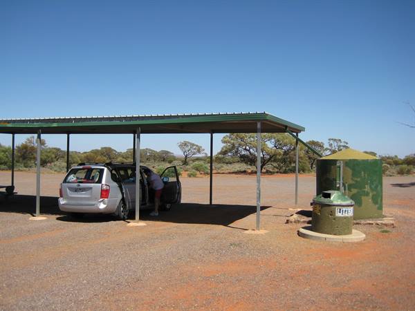 Car parked under a metal shelter in the outback, green water tank nearby