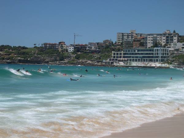 Surfers on turquoise waves at Bondi Beach, buildings along the coast