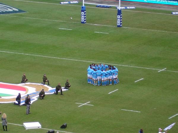 Blue rugby team huddling on the grass pitch, photographers near the logo