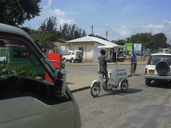 Man pedals an ice cream tricycle along a busy road