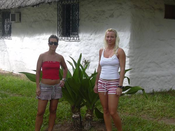 Two women in summer tops beside a green plant in front of a white house