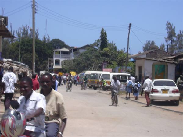 Motorcyclist in foreground, cyclists and minibuses on dirt road among buildings and trees