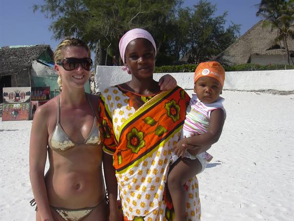 Tourist in bikini with woman in orange kanga holding baby on white sand beach