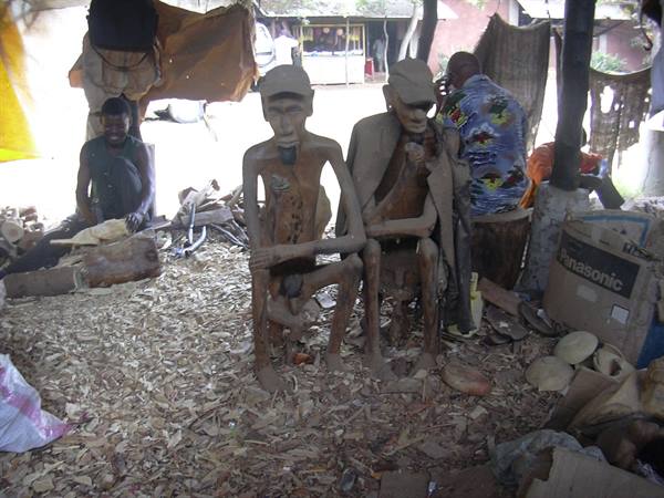 Carver beside two seated wooden statues in a wood-shaving covered workshop