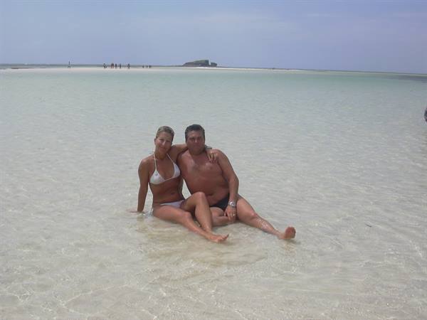 Couple sitting in shallow clear water on white sandbank under bright sky