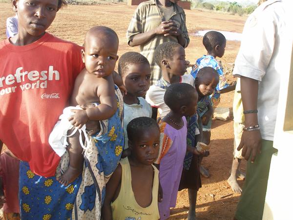 Children queueing on red dirt, a girl in a red Coca-Cola shirt carries a baby