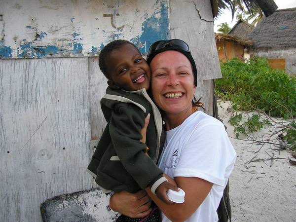 Woman hugging a smiling child in front of a weathered wooden hut