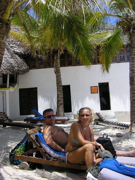 Couple in swimwear on wooden loungers under palm trees, white sand beach