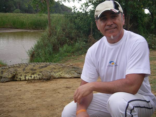 Man crouching next to a large crocodile on a sandy riverbank