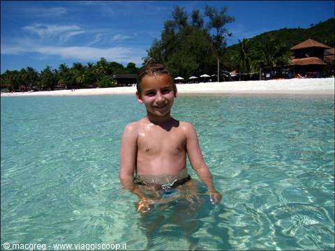 Smiling boy in turquoise water in front of white beach and palms