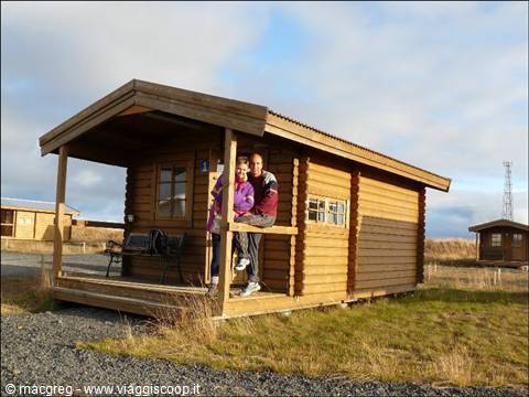 Smiling couple on the porch of a wooden cabin, campground under cloudy sky