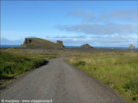 Gravel road through grassy fields toward rocky coast and blue sea
