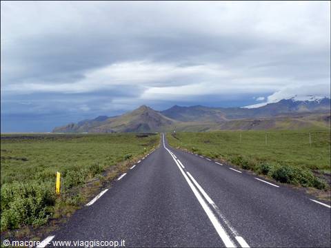 Straight asphalt road across green plains, volcanic mountains and cloudy sky