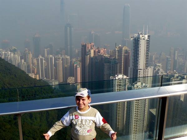Boy in cap on glass terrace at the Peak, Hong Kong skyscrapers in haze