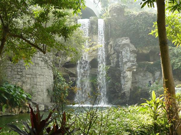 Artificial waterfall over rocks and stone walls, surrounded by lush tropical plants
