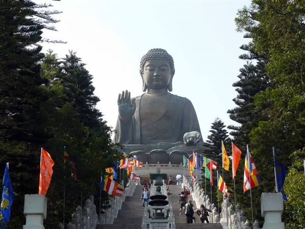 Large bronze Buddha statue among trees, visitors climbing steps with colorful flags, daylight