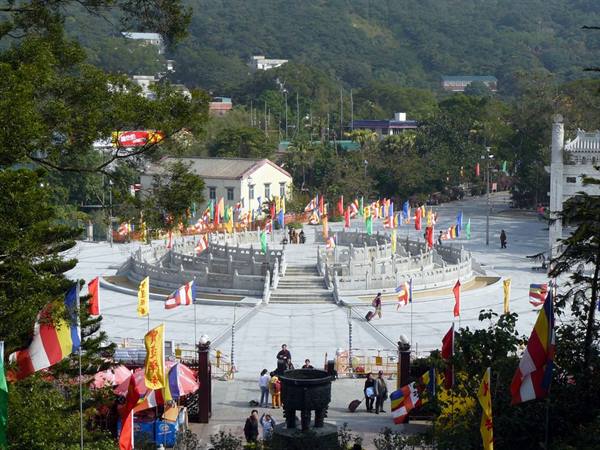 Circular stone plaza with colorful prayer flags among trees, daylight, visitors walking