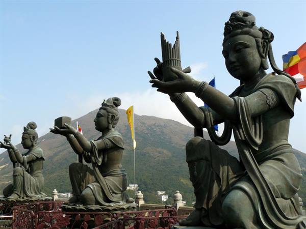Bronze bodhisattva statues worshiping before green mountain, daylight, colorful flags waving