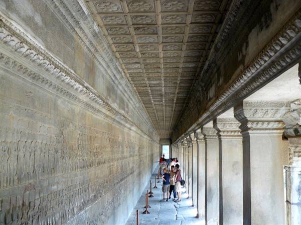 Stone gallery with carved bas-reliefs and pillars, visitors admire the Angkor Wat reliefs