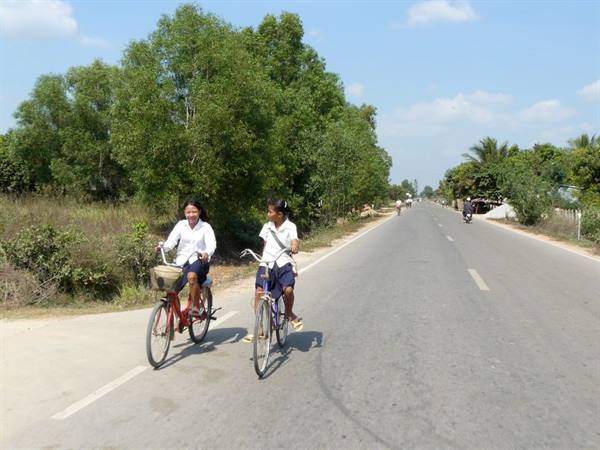 Two girls in white uniforms ride bicycles along a sunny asphalt road