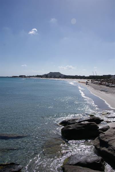 Wet rocks and sandy beach under bright sun, hill in the background