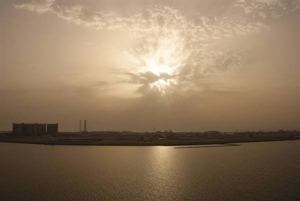 Sun breaking through clouds over calm bay, industrial skyline and towers on shore