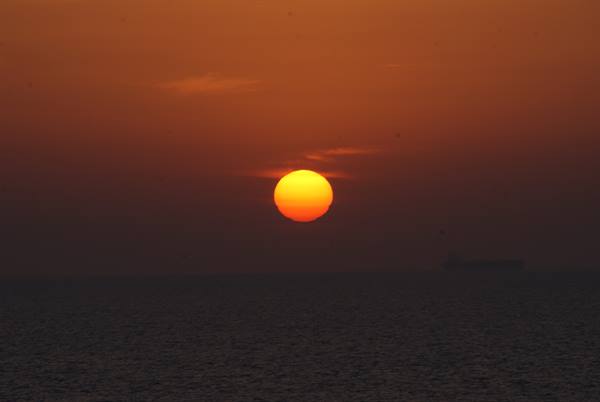 Orange sun low over dark sea, a cargo ship fading in the haze